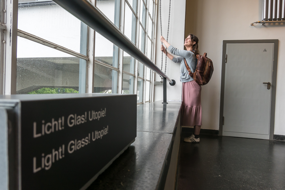 Eine Frau an einem großen Fenster. Im Vordergrund ein Schriftzug: Licht! Glas! Utopie!